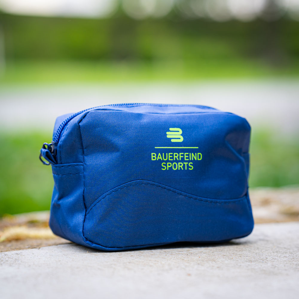 Eine kleine, blaue Kosmetiktasche von Bauerfeind Sports mit Reißverschluss liegt auf einem Betonuntergrund im Freien. Die Tasche zeigt das Bauerfeind-Logo und ist kompakt für den Outdoor-Einsatz.