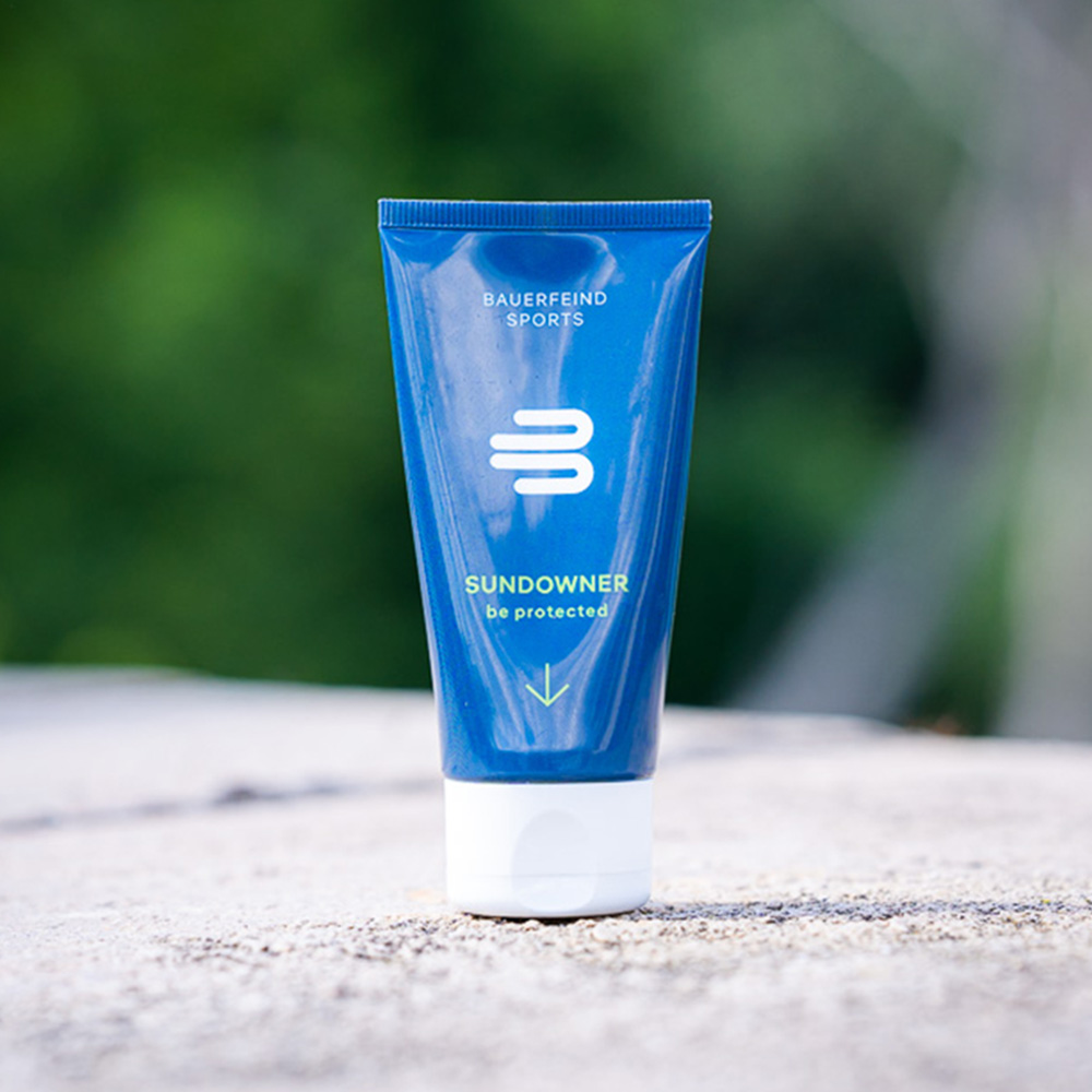 Eine blaue Tube der Bauerfeind Sports Sundowner Sonnencreme mit LSF 30 steht auf einem Stein, umgeben von einer grünen Naturkulisse. Die Sonnencreme bietet wasserfesten und veganen Sonnenschutz für Aktivitäten im Freien.