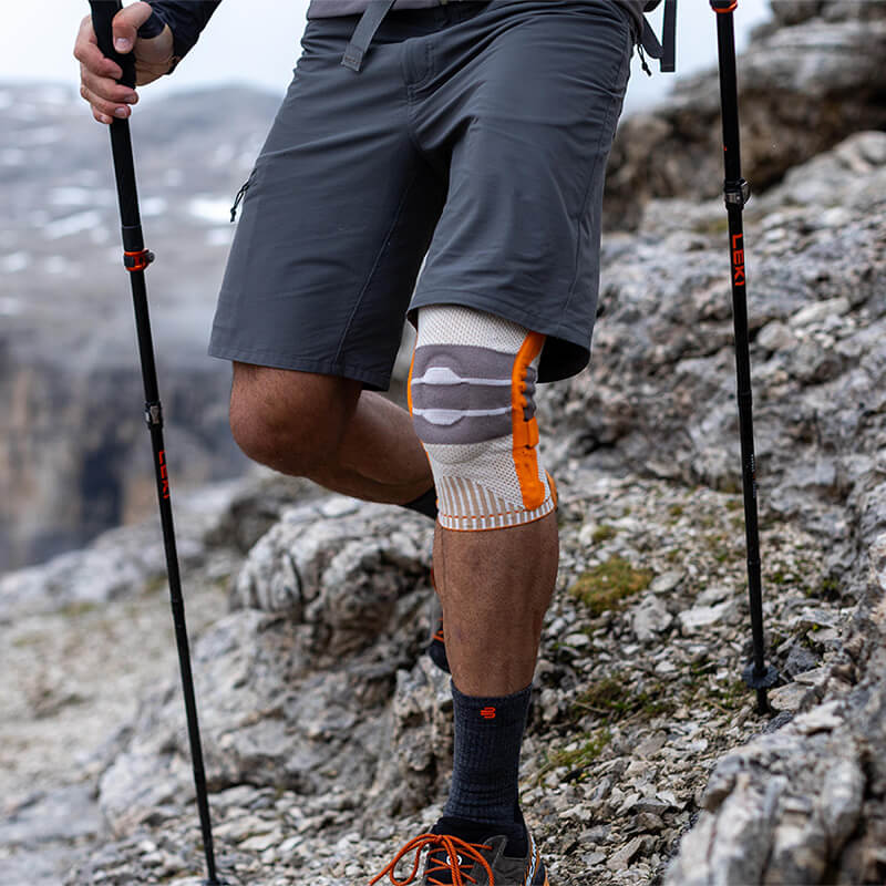 Mann beim Tragen der Wander-Kniebandage in den Bergen beim Wandern mit Stöcken in felsigem Terrain.