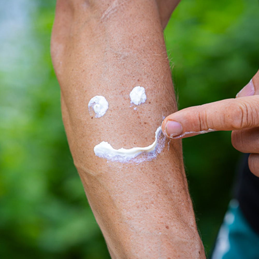 Auf einem Unterarm ist Sonnenschutzcreme von Bauerfeind Sports aufgetragen, die ein lächelndes Gesicht bildet. Eine Hand zeigt auf den "Mund" des Smileys. Die Szene findet im Freien statt, umgeben von grüner Natur.