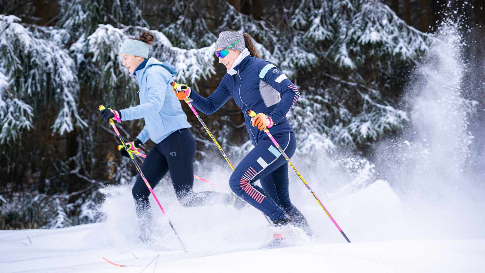 Zwei Frauen beim Skilanglauf durch verschneite Wälder. Sie tragen farbenfrohe Wintersportbekleidung und gleiten dynamisch durch den Schnee. Die Szenerie ist geprägt von dicht verschneiten Tannen, die das Winterwunderland unterstreichen.