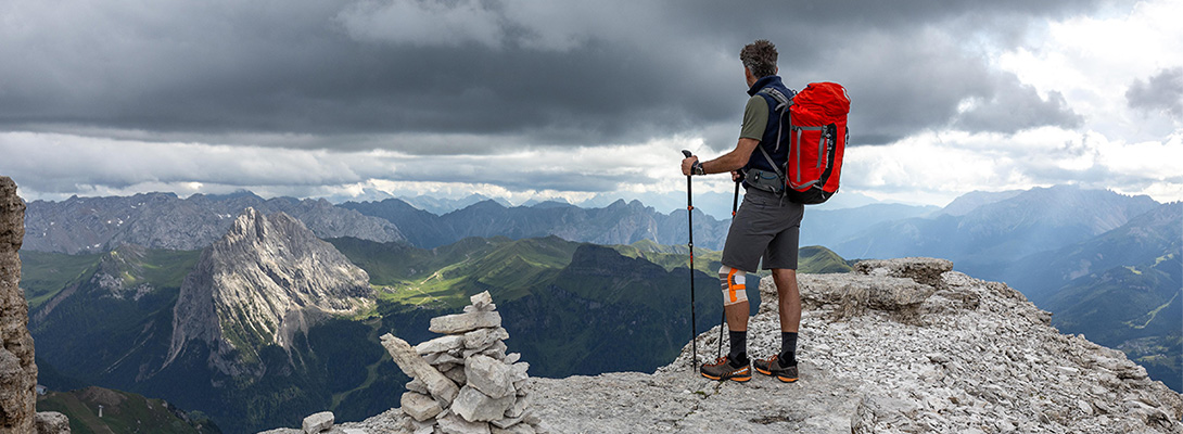 Bergsteiger mit rotem Rucksack und Wanderstöcken auf einer Bergspitze, er trägt eine Wander Kniebandage und blickt in die Ferne