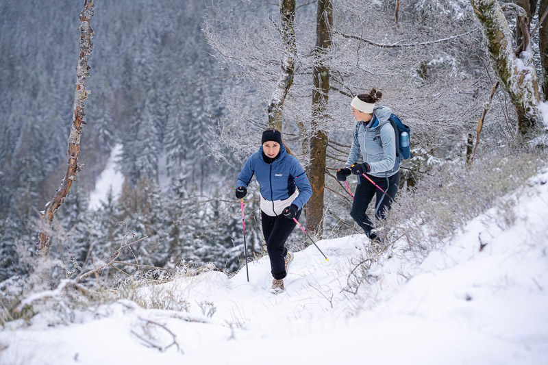 Zwei Frauen wandern mit Trekkingstöcken durch eine verschneite Winterlandschaft. Sie tragen warme Funktionskleidung, eine blaue Jacke und eine hellblaue Jacke, sowie Handschuhe und Stirnbänder.