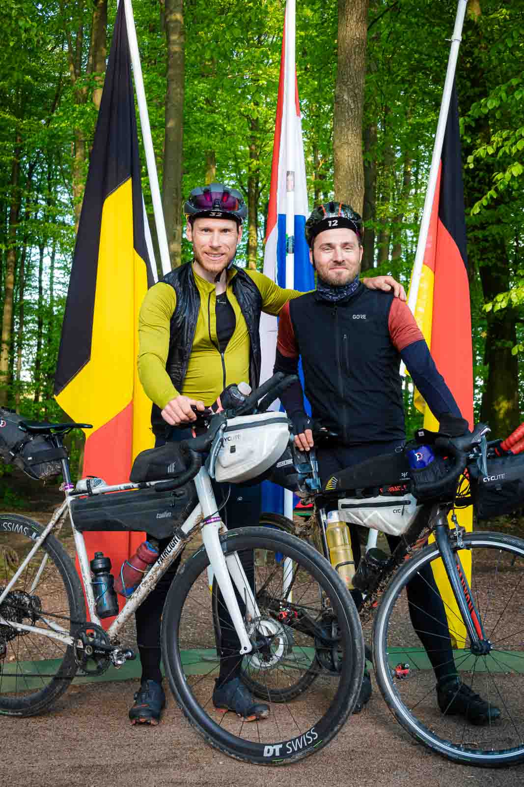 Am Dreiländereck von Deutschland, Belgien und den Niederlanden stehen zwei Radfahrer und lachen in die Kamera.