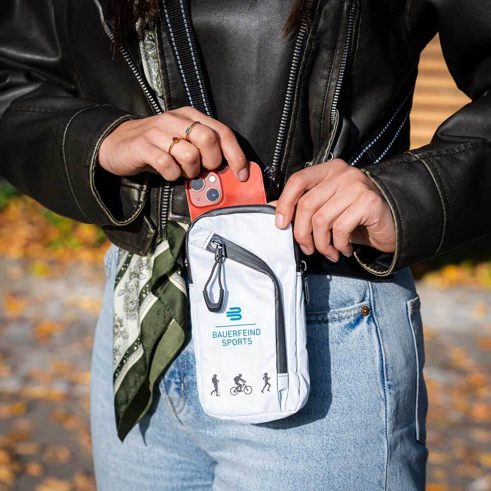 Eine Person verstaut ein Smartphone in der grauen Bauerfeind Sports Tasche, die an der Hüfte getragen wird. Die Tasche ist kompakt und eignet sich perfekt für Outdoor-Aktivitäten.