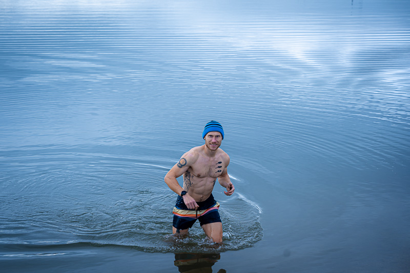 Ein Mann watet aus dem See, während er beim Eisbaden in Badehose und mit blauer Mütze in der herbstlichen Umgebung fotografiert wird.