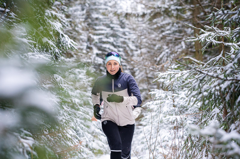 Eine Läuferin joggt durch einen verschneiten Waldweg. Die Bäume um sie herum sind dicht mit Schnee bedeckt. Sie trägt eine hellgraue Laufjacke mit schwarzen Akzenten, eine schwarze Hose und eine blaue Stirnbinde mit Sonnenbrille.